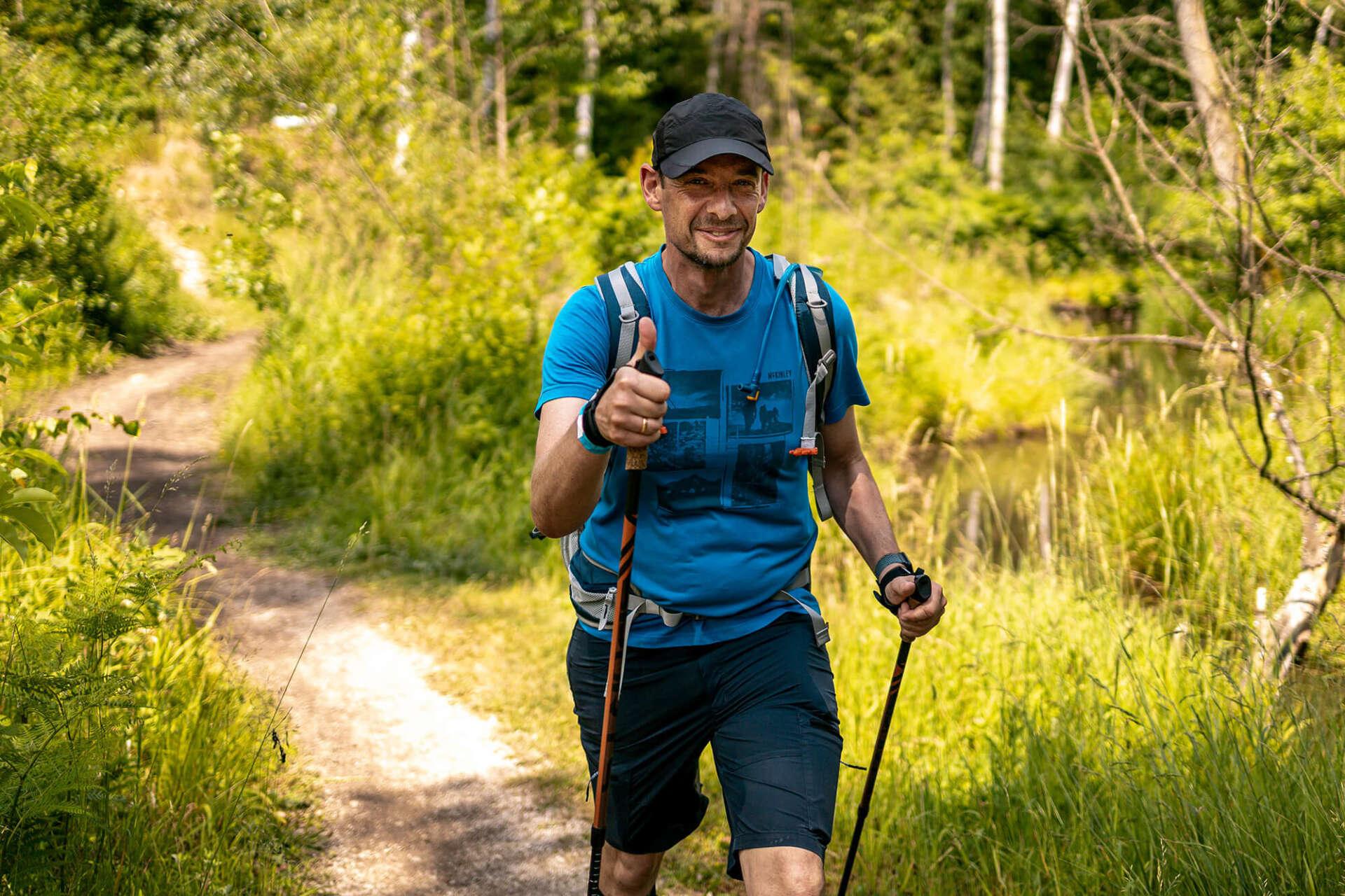 Man med ryggsäck och vandringsstavar vandrar längs en skogsstig och gör tummen upp mot kameran.
