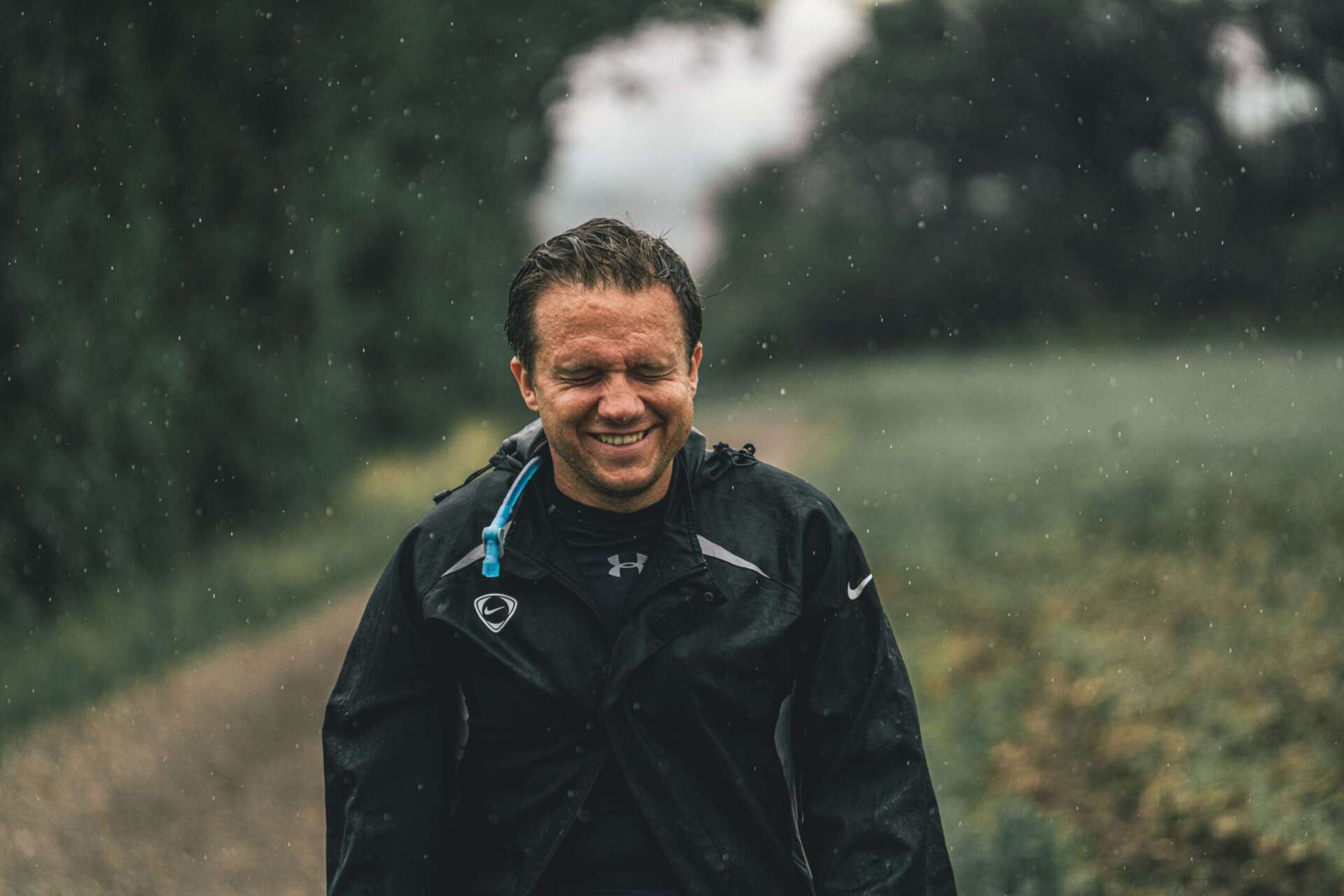En leende vandrare i svart regnjacka på en stig i naturen medan regnet faller.